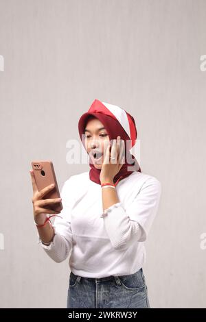 Die junge asiatische Frau mit rotem Hijab ist überrascht mit ihrem Handy in der Hand, indonesien Unabhängigkeitstag Konzept Stockfoto