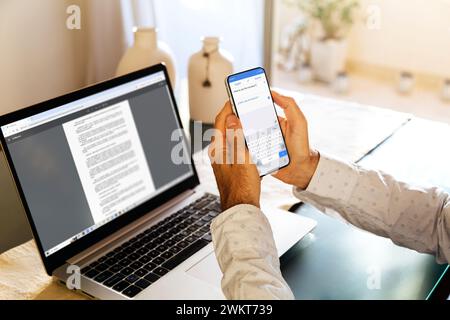 Mann, der die Google Translate-Anwendung in einem Smartphone verwendet. Er ist zu Hause und liest ein digitales Buch in einem Laptop. Mobiltelefon mit übersetzter App für Sprache Stockfoto