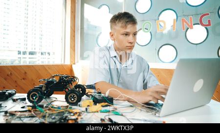 Intelligenter Schüler, der an einem Laptop arbeitet, während er die Konstruktion eines Fahrzeugmodells analysiert. Glücklicher Highschool-Junge, der Computer benutzt, um zu lernen oder mehr über Robotermodelle zu lernen Stockfoto