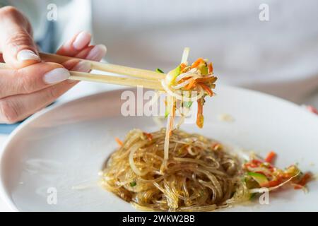 Eine Frau mit Essstäbchen in der Hand nimmt gebratene Glasnudeln mit Gemüse, Japchae (auch Chapchae oder 잡채 auf Koreanisch) ist ein beliebter koreanischer Di Stockfoto