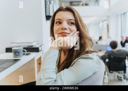 Kontemplative junge Frau im Büroumfeld mit Kollegen im Hintergrund. Sie lächelt, wenn sie etwas ansieht. Stockfoto