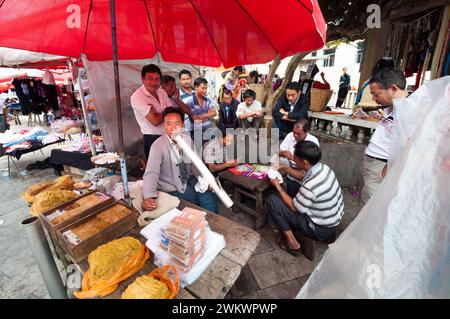 Ein Mann, der eine Triditionspfeife raucht, umgeben von einer Gruppe Männer, die Karten spielen, an einem Verkaufsstand im ländlichen Yunnan China Stockfoto