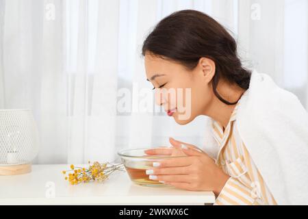 Junge asiatische Frau, die am Tisch im Schlafzimmer Dampf einatmet Stockfoto