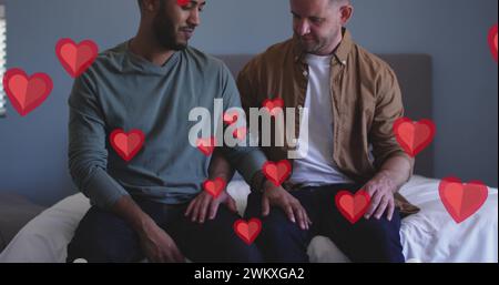 Bild von roten Herzen über verschiedenen männlichen Paaren, die auf dem Bett sitzen Stockfoto