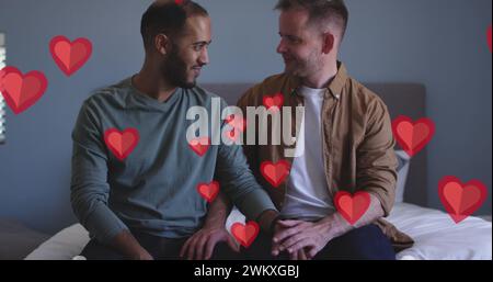 Bild von roten Herzen über verschiedenen männlichen Paaren, die auf dem Bett sitzen Stockfoto