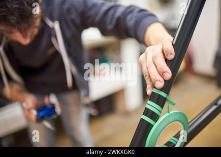 Unerkennbarer Mann, der einen Fahrradrahmen mit Abdeckband für ein individuelles Maldesign in seiner Fahrradwerkstatt vorbereitet, ein handgefertigter kreativer Prozess. Stockfoto