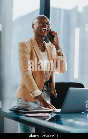 Eine afrikanische Geschäftsfrau in einem Firmenbüro verwendet ihr Smartphone, um über ihre Arbeit zu sprechen und zu diskutieren. Sie steht mit einem Laptop neben ihrem Schreibtisch und zeigt Professiona Stockfoto