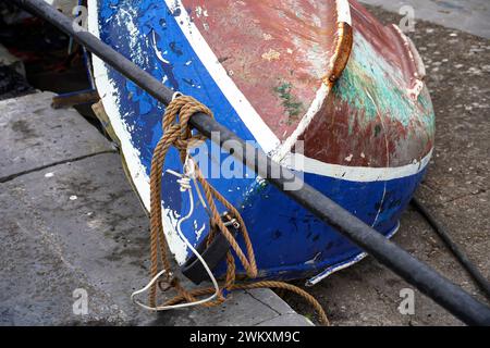 Ein kleines, umgedrehtes Ruderboot mit abblätternder Farbe, Moos und rostenden Armaturen liegt unangenehm auf einem blauen Yachthafen. Stockfoto