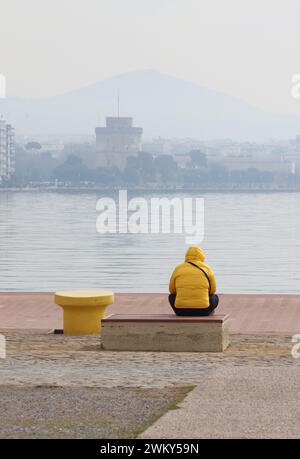 Ruhig sitzen. Rückansicht Perspektive. Mann in der gelben Jacke, der auf den weißen Turm im nebeligen Hintergrund blickt und sich an der ägäis entspannt. Stockfoto