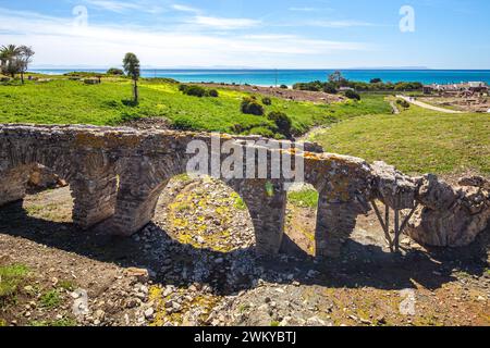 Ruinen einer alten Brücke in einem grünen Feld, mit dem blauen Meer und dem klaren Himmel im Hintergrund. Die Ruinen stammen von der römischen Stätte von Baelo Claudia, Lo Stockfoto