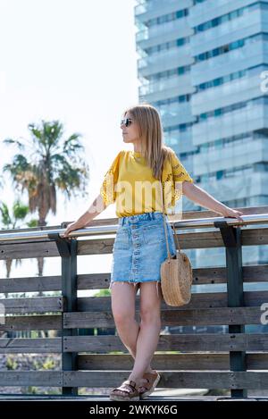 Eine junge Frau blickt in die Ferne, lehnt sich auf ein Geländer mit einer Kulisse aus Palmen und modernen Gebäuden unter einem klaren blauen Himmel. Sie trägt einen Stockfoto