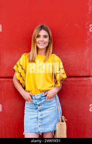 In diesem Porträt steht eine junge Frau selbstbewusst vor einer satten roten Wand, ihre gelbe Bluse prallt vor Farbe und Energie. Der Jeansrock sorgt für eine besondere Note Stockfoto