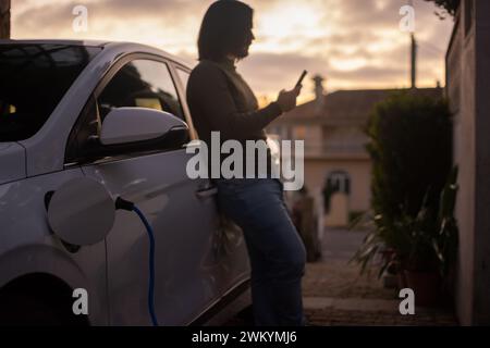 Ein Mann lädt ein Elektroauto, das im Hausbereich geparkt ist, und stellt eine EV-App auf dem Smartphone ein. Konzept der grünen Energie und der Nachhaltigkeit Stockfoto