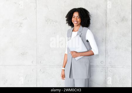 Eine afroamerikanische Frau strahlt Professionalität aus und hält sich selbstbewusst mit einem Laptop in der Hand, in einem Business-Casual-Outfit, das moderne Arbeitskleidung mit Komfort perfekt verbindet Stockfoto