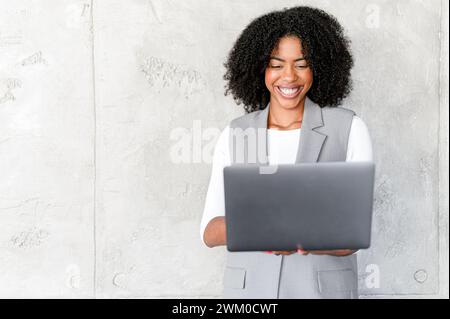 Eine fröhliche afroamerikanische Geschäftsfrau hält einen Laptop, ihr ansteckendes Lächeln deutet auf Produktivität und Benutzerfreundlichkeit in der Unternehmensumgebung hin. Stockfoto