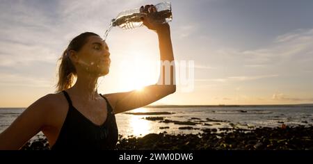 Sportliche Frau, die erfrischendes Wasser aus der Flasche ins Gesicht gießt, nach dem Fitness-Workout im Freien am Strand Stockfoto