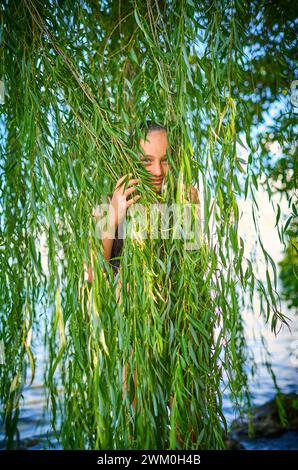 Verspieltes Mädchen, das sich hinter Blättern von Weidenbäumen versteckt Stockfoto