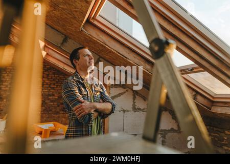 Lächelnder Mann mit überkreuzten Armen, der zu Hause aus dem Fenster schaut Stockfoto