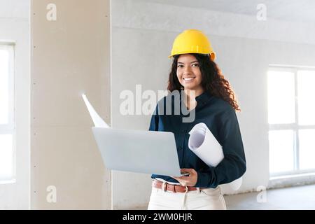 Ein lächelnder junger Architekt hält Laptop und Blaupausen vor Ort Stockfoto