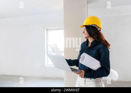 Ein lächelnder Architekt hält einen Laptop in der Hand und untersucht den Standort mit Blaupausen Stockfoto