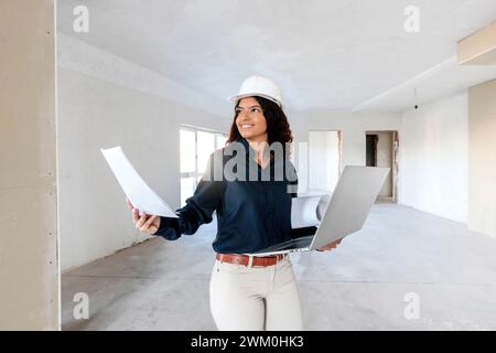 Ein lächelnder junger Architekt hält einen Laptop und untersucht die Baustelle mit Blaupausen Stockfoto