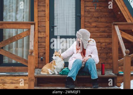Glückliche Frau, die mit Corgi-Hund auf der Veranda einer Blockhütte sitzt Stockfoto