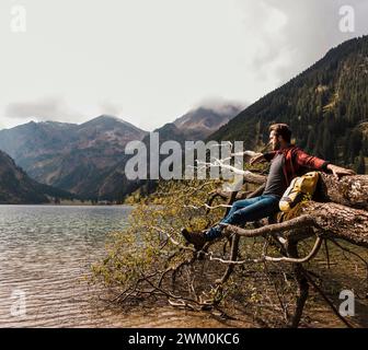 Junger Mann, der auf einem umgestürzten Baumstamm am Vilsalpsee und in den Bergen in Tirol sitzt Stockfoto