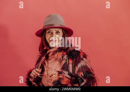 Glückliche Seniorin, die eine Pelzjacke vor der roten Wand trägt Stockfoto