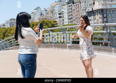 Junge lateinfrau, die draußen mit ihrem Telefon steht und eine Geigerin filmt, die Geige für soziale Medien spielt. Stockfoto