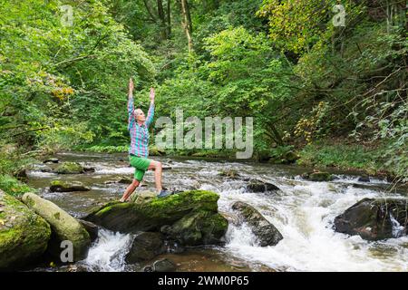 Ältere Frau, die mit erhobenen Händen im Wald um Bach trainiert Stockfoto