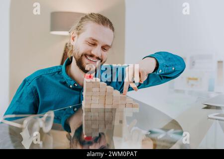 Glücklicher Geschäftsmann, der auf Stufen aus Holzspielblöcken am Schreibtisch im Büro spielt Stockfoto