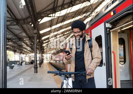 Ein lächelnder Mann, der das Handy benutzt und am Bahnhof in der Nähe des Bahnhofs am Fahrrad steht Stockfoto