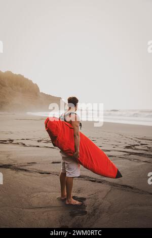 Junger Mann, der Surfbrett trägt und am Strand steht Stockfoto