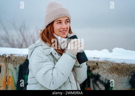 Lächelnde Frau trinkt Tee aus Thermoskanne in der Nähe der Wand im Winter Stockfoto