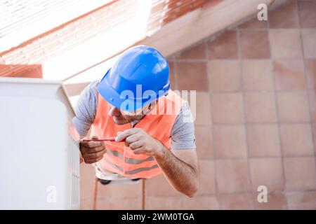 Techniker mit blauem Schutzhelm, der die Klimaanlage im Haus repariert, das gerade renoviert wird Stockfoto
