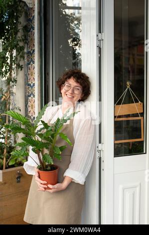 Glücklicher Botaniker, der die Pflanze hält und sich an die Ladentür lehnt Stockfoto
