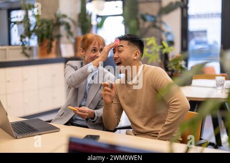 Verspielte, reife Geschäftsfrau, die im Büro einen Haftnotiz auf die Stirn des Kollegen klebt Stockfoto