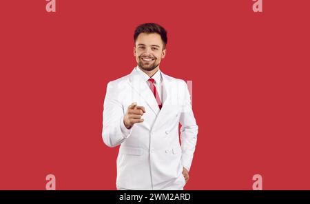 Glücklicher, gutaussehender junger Mann in einem modischen weißen Anzug, der lächelt und mit dem Finger auf dich zeigt Stockfoto