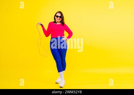 Hübsch lächelnde, pralle Frau in sportlichem Oberteil und Leggings, die ein Springseil in den Händen hält Stockfoto