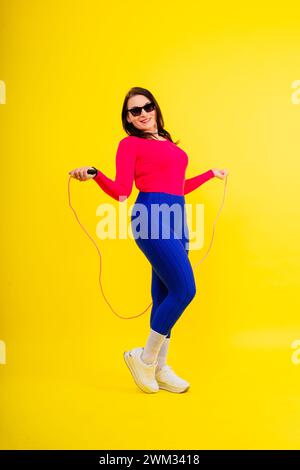 Hübsch lächelnde, pralle Frau in sportlichem Oberteil und Leggings, die ein Springseil in den Händen hält Stockfoto