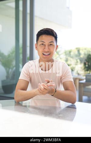 Ein junger asiatischer Mann sitzt an einem Tisch und lächelt selbstbewusst bei einem Videoanruf Stockfoto