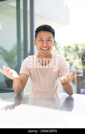 Ein junger asiatischer Mann gibt Gesten, während er an einem Tisch sitzt, während er ein Videogespräch führt Stockfoto