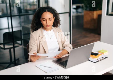 Ernsthafte selbstbewusste brasilianische oder hispanische Geschäftsfrau mit lockigem Haar, ceo des Unternehmens, Finanzmanager, sitzt am Schreibtisch in einem modernen Büro, arbeitet in einem Laptop, konzentriert sich auf das Studium von Finanzdokumenten Stockfoto