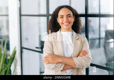 Schöne erfolgreiche lateinische junge Geschäftsfrau mit lockigen Haaren in eleganter Kleidung, Corporate Marketing Manager, ceo des Unternehmens, stehend im modernen Büro mit überkreuzten Armen, Blick in die Kamera, lächelt Stockfoto