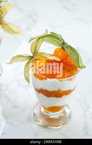 Blick von oben auf das mehrschichtige Dessert mit Proteinjoghurt und Physalis-Früchten in einem Glasgefäß auf Marmortisch Stockfoto