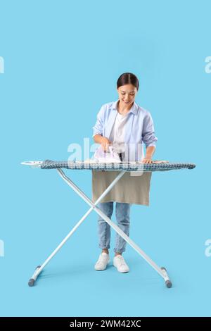 Junge asiatische Frau, die Kleidung auf blauem Hintergrund bügelt Stockfoto