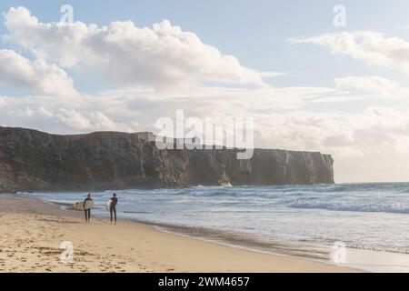 Zwei Surfer am Strand Praia do Tonel mit Brettern vor den Klippen an der atlantikküste mit Fortaleza de Sagres, Sagres, Algarve, Portugal Stockfoto