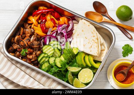 Vegane Fajitas mit Pilzen, Paprika, rote Zwiebeln, Gurke, Avocado und Koriander in der Pfanne mit glutenfreien Tortillas auf weißem Holztisch mit Stockfoto