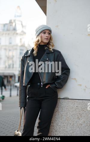 Stilvolles, schönes Mädchen-Model in modischer Kleidung mit Hut, schwarzer Lederjacke, Pullover und Hose steht neben einer Wand auf der Straße Stockfoto