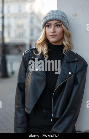 Urbanes modisches, hübsches, schickes Mädchen mit blondem Haar und einem Hut in schwarzer, lässiger Rock-Kleidung mit Lederjacke und Pullover in der Stadt. Niedliche Frau Stockfoto